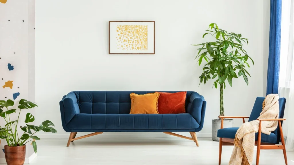 Mid-century modern chair with a blanket and a large sofa with colorful cushions in a spacious living room interior with green plants and white walls.
